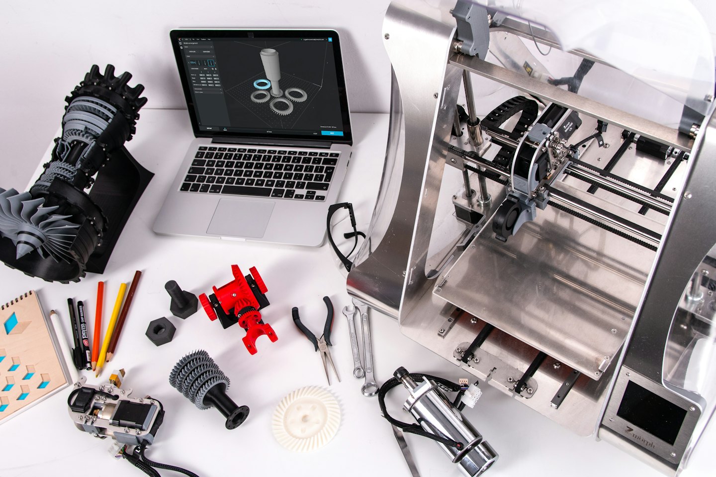 Laptop und 3D-Drucker auf einem weißen Schreibtisch mit Prototypen.
